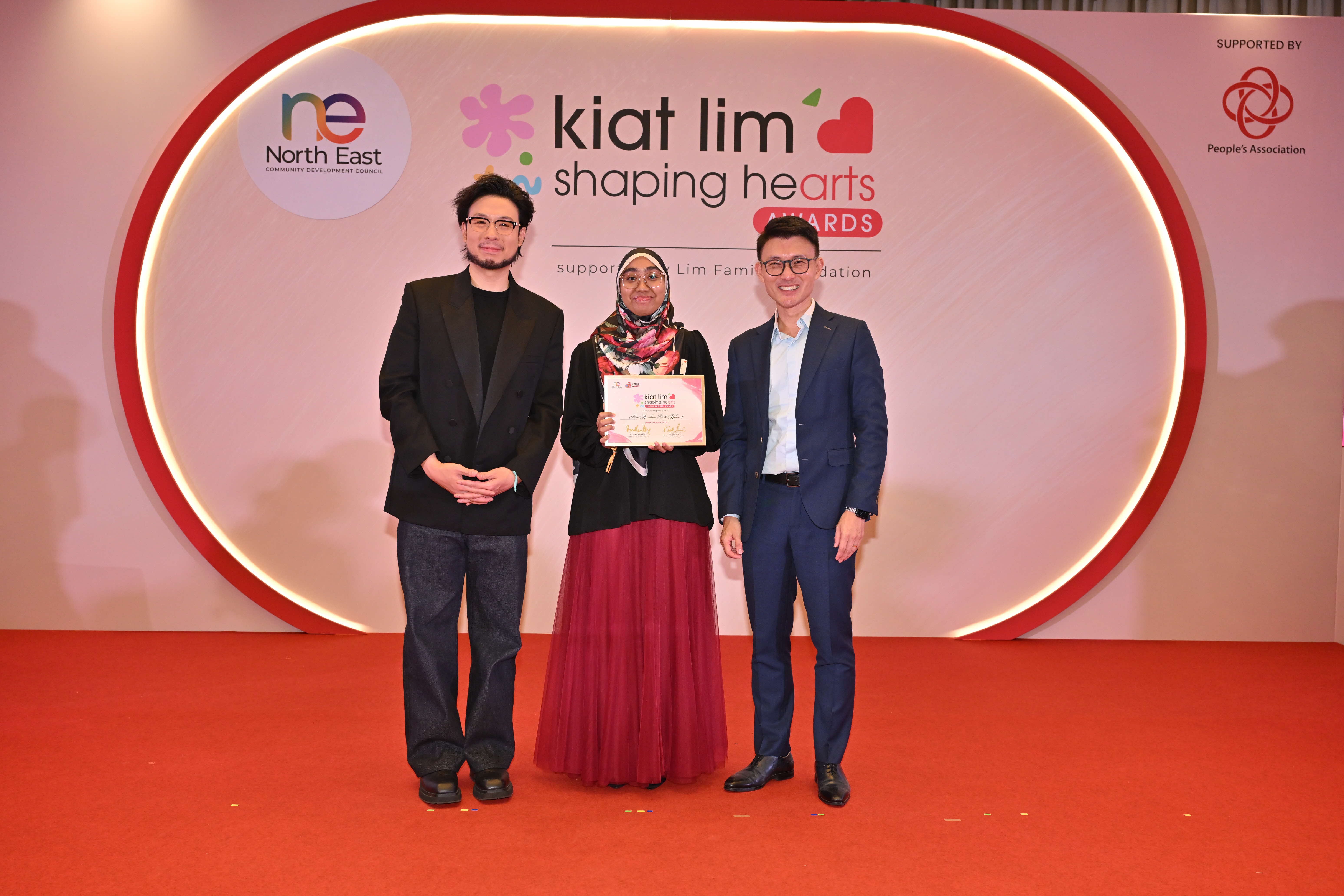 Award recipient standing on stage with the presenter, holding their certificate and posing for a group photo during the award ceremony, with a backdrop reading ‘Kiat Lim-Shaping Hearts Awards'.